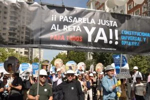 Crowd of protesters marching under a large banner that reads '¡ PASARELA JUSTA AL RETA YA!'; participants wear hard hats and carry signs.
