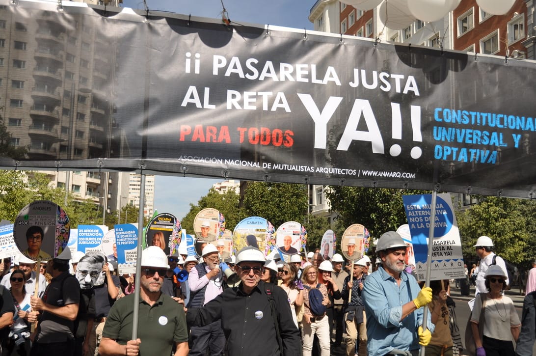 Crowd of protesters marching under a large banner that reads '¡ PASARELA JUSTA AL RETA YA!'; participants wear hard hats and carry signs.