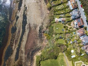 773632 - InmoCasas y Lares | C. Abogados News Aerial view of a rocky shoreline on the left and a row of houses with green gardens on the right, seen from above by the coast.