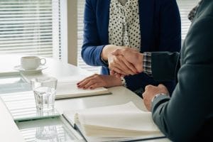 773732 - Briones Abogados | C. Abogados News Two people in business attire shake hands across a white table during a meeting, with open notebooks and a glass of water nearby.