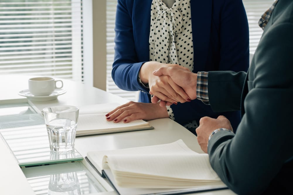 Two people in business attire shake hands across a white table during a meeting, with open notebooks and a glass of water nearby.