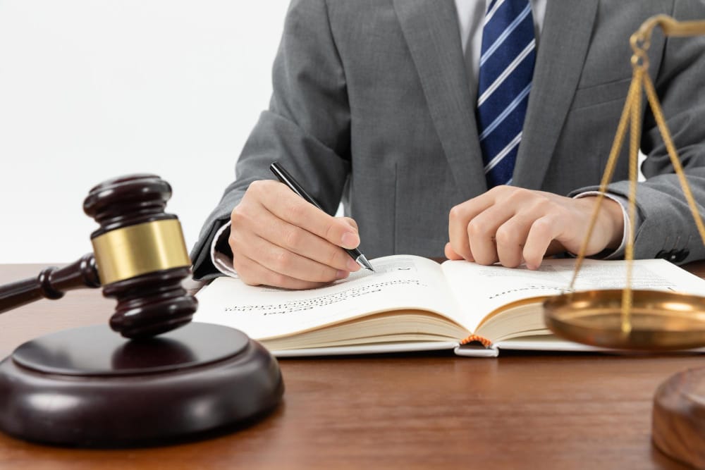 Judge's gavel and scales of justice sit on a desk as a lawyer in a gray suit writes in an open law book.
