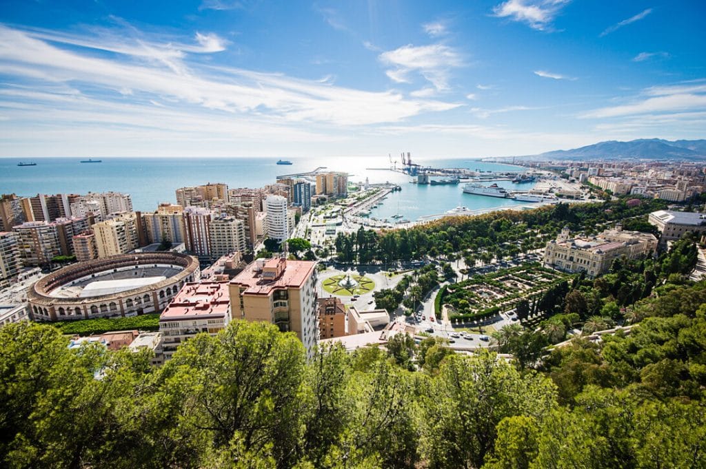 Aerial view of a sunny coastal city with a harbor, ships, and a circular stadium among buildings near the water.