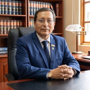 Man in a blue suit seated at a desk in an office with wooden bookshelves behind him.