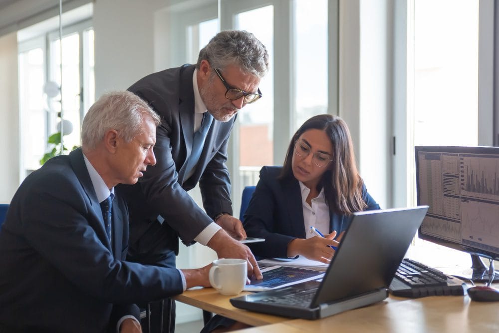 Three professionals in suits collaborate around a laptop at a bright office desk.
