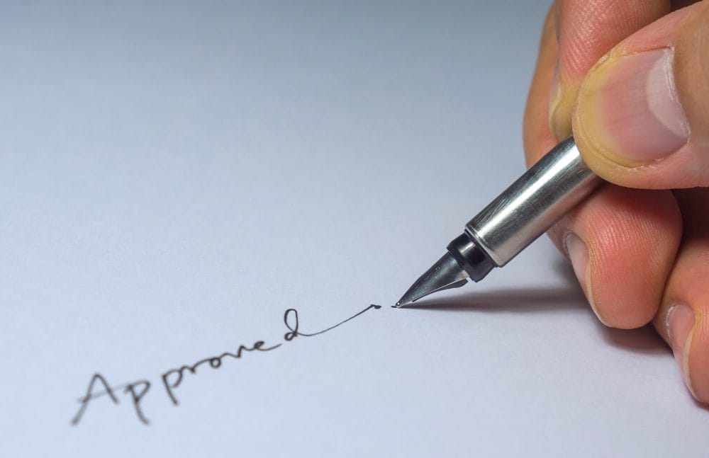 Close-up of hand writing 'Approved' with a silver pen on white paper.