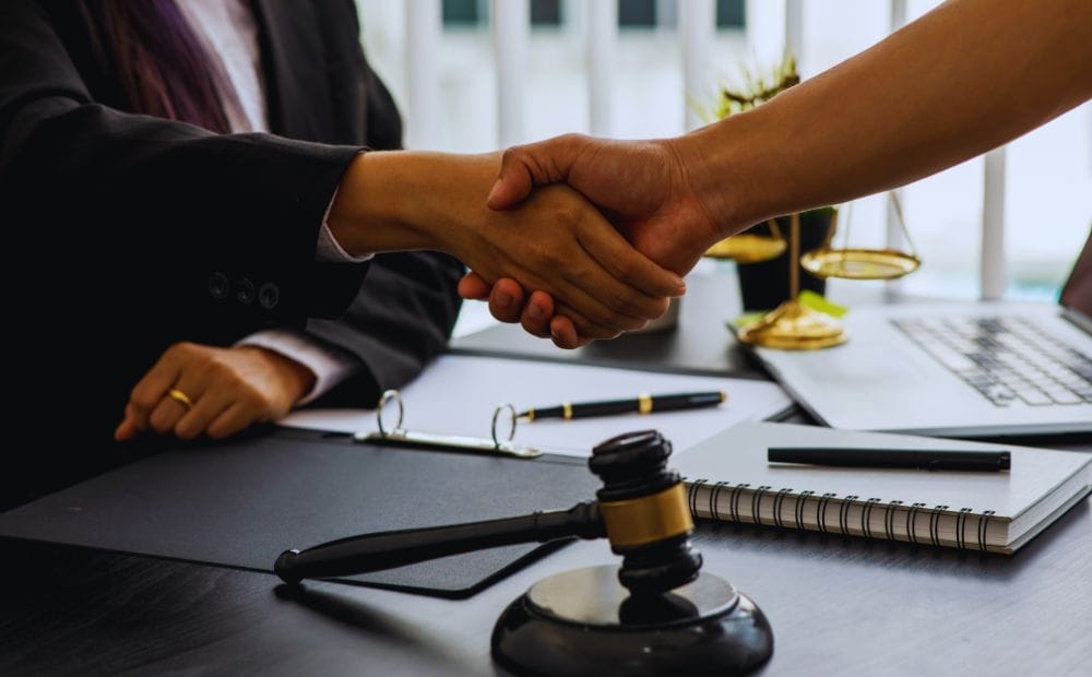 Two professionals shake hands over a desk in a law office, with a gavel and notebook in view